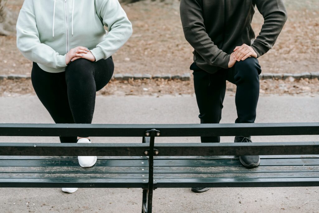 Crop faceless fit man and overweight woman putting legs on wooden bench while training together in autumn park