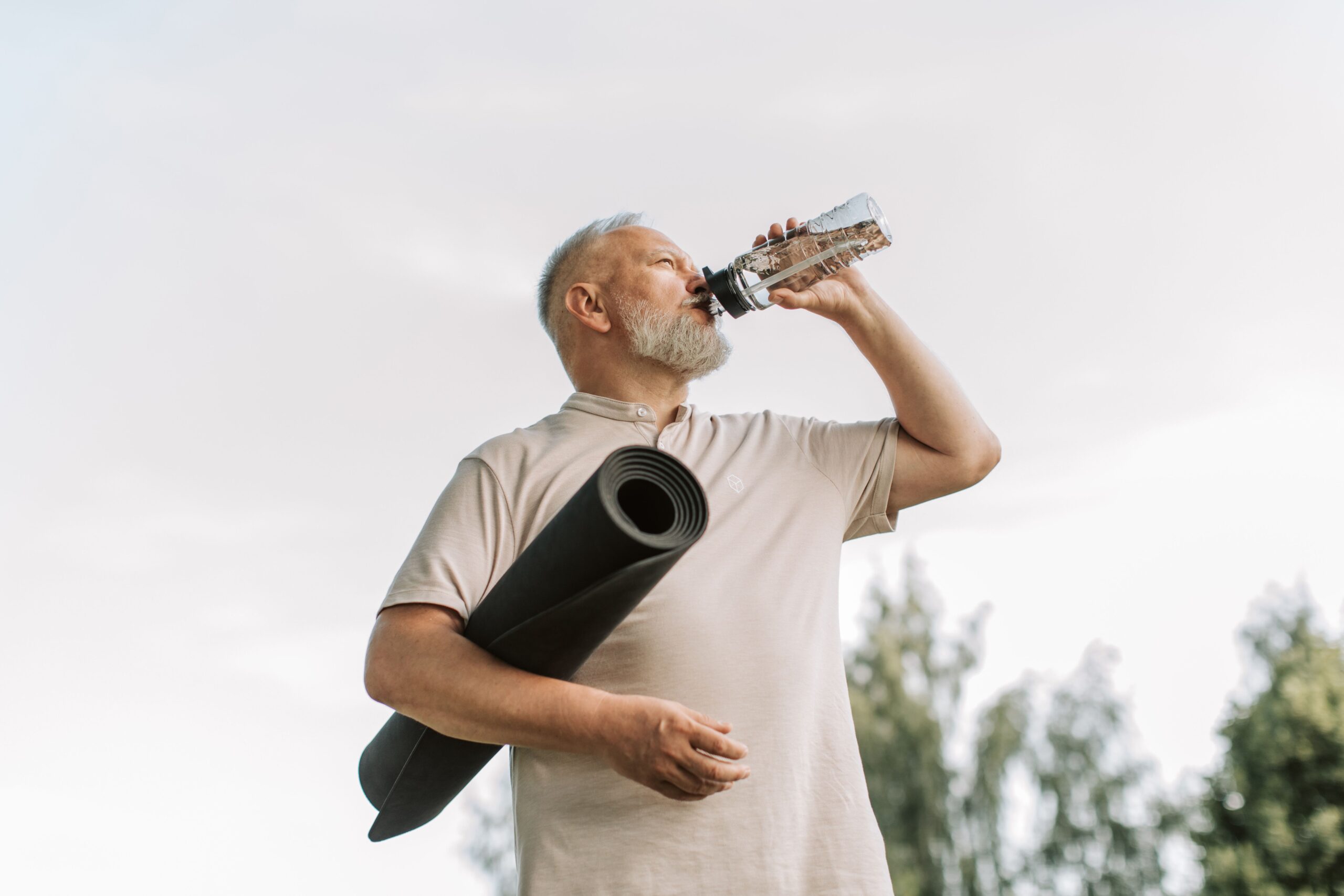 A senior man, drinking water from a water bottle, outdoors, with rolled up yoga mat under one arm.