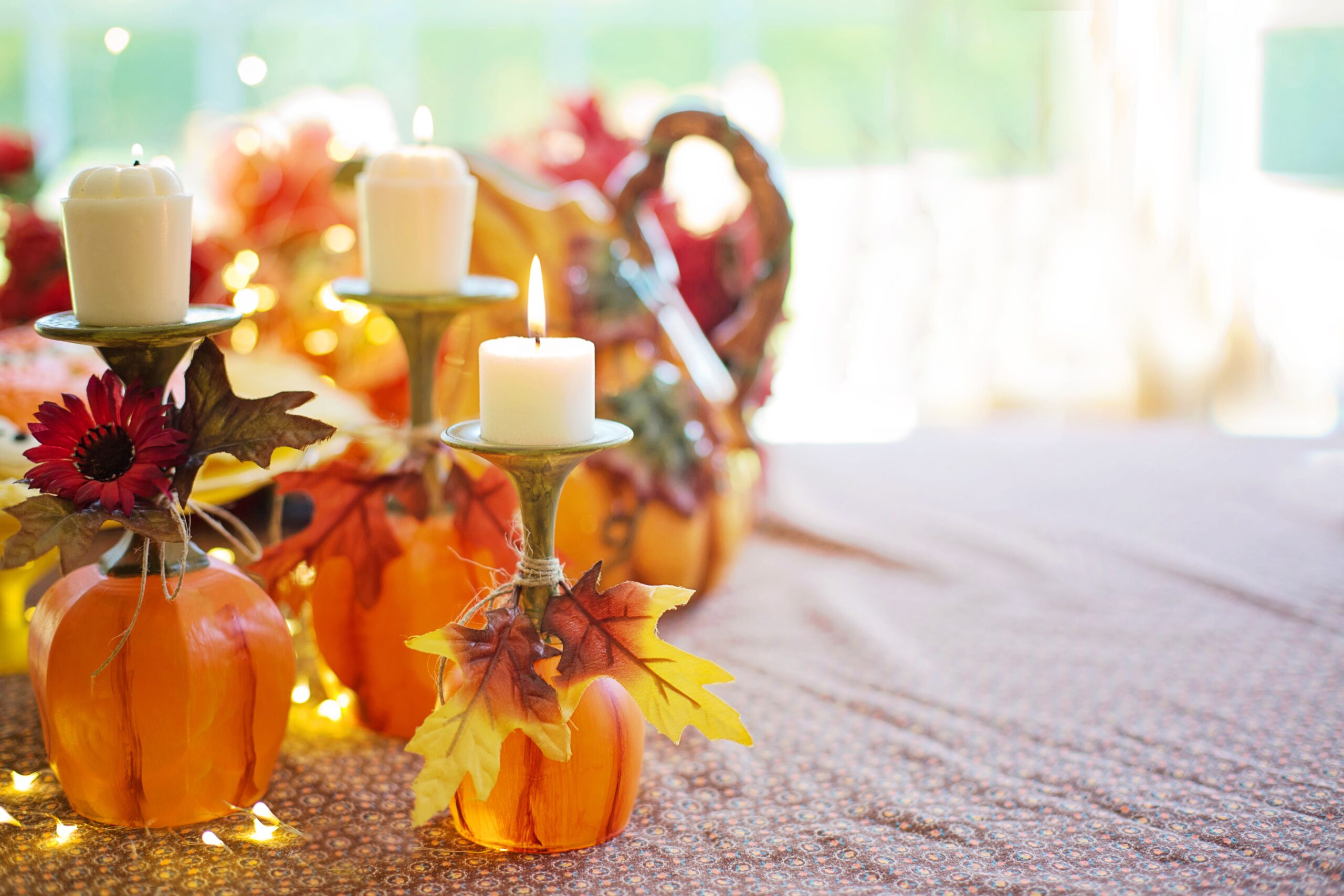 DIY Thanksgiving centerpiece of upcycled wine glasses painted orange like pumpkins and used as candle holders, decorated with twine, silk maple leaves, and a red daisy, with warm string lights and a woven cornucopia in the soft-focus background.