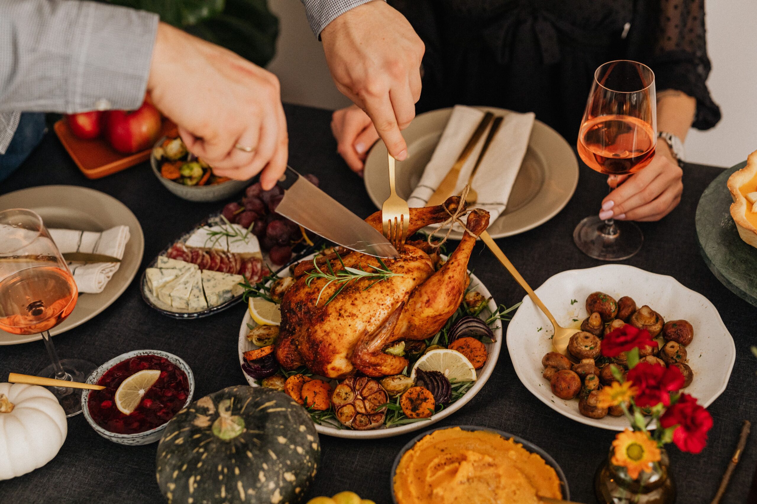 Overhead view of two people carving a roasted turkey at a Thanksgiving dinner table surrounded by side dishes — cranberry sauce, mashed sweet potatoes, sautéed mushrooms, roasted vegetables, a cheese and charcuterie board, grapes, and glasses of rosé wine.