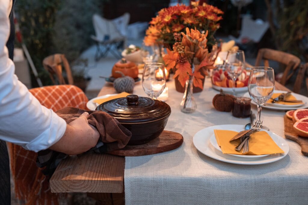 Person in a white shirt setting a covered brown stoneware casserole dish onto a wooden board at an outdoor Thanksgiving table, styled with a linen tablecloth, gold napkins, wine glasses, lit candles, orange chrysanthemums, and sliced grapefruit at dusk.