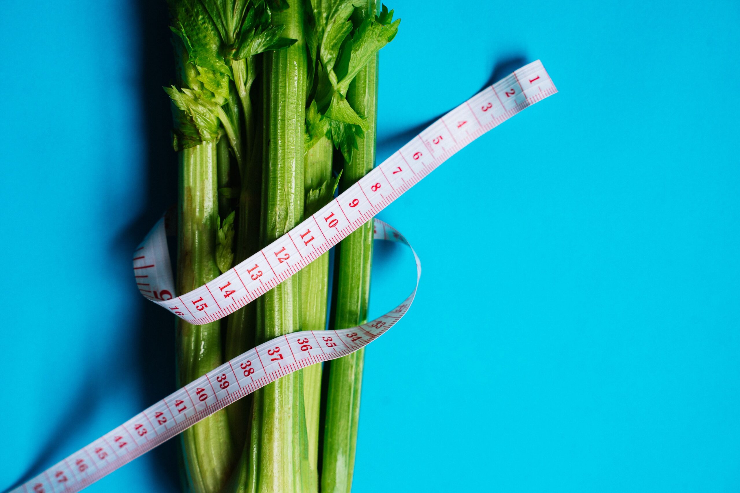 Celery stalks wrapped in a white measuring tape against a blue background, representing dietary changes paired with weight loss drugs.