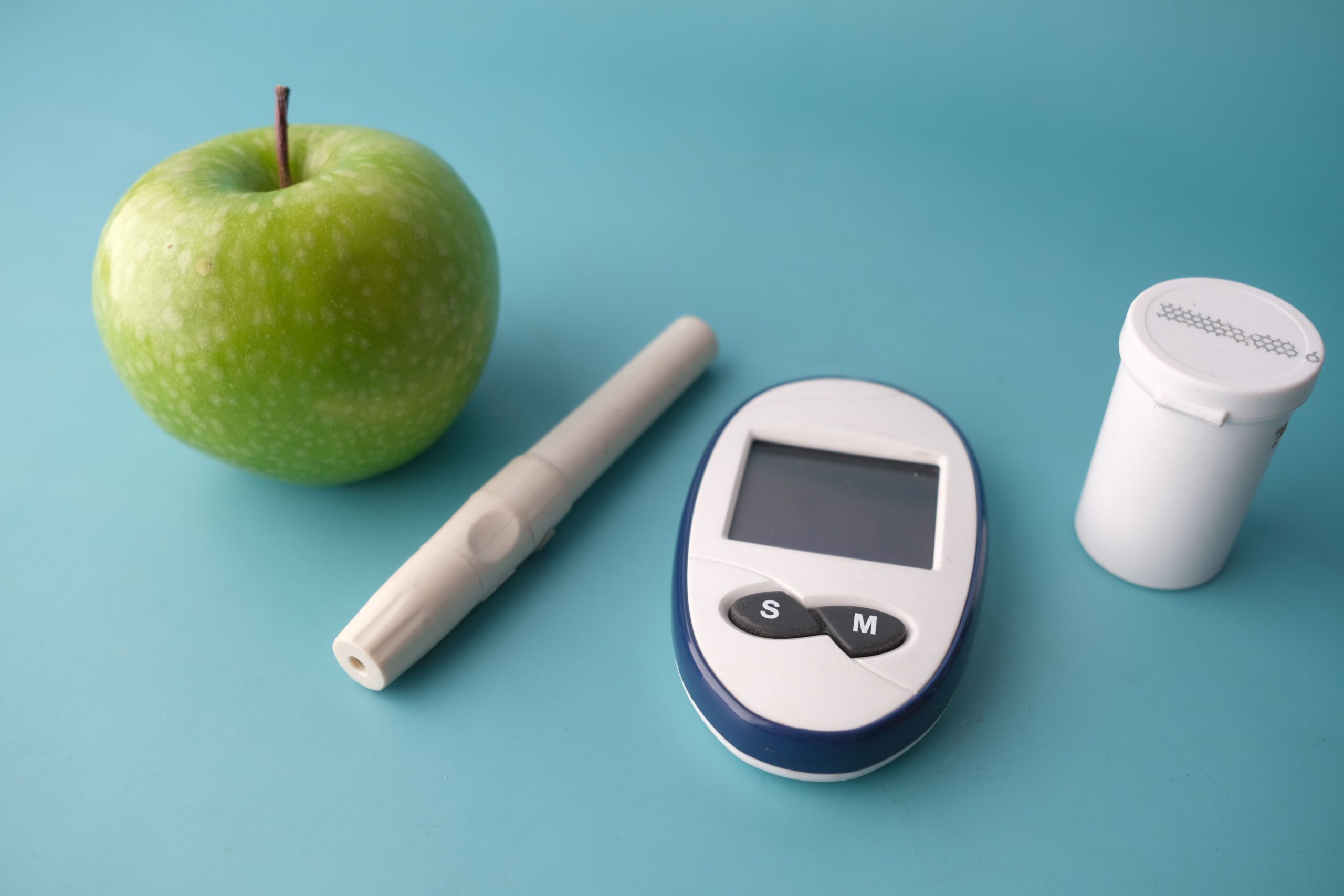 Green apple, lancet pen, glucose meter and test strip vial on a blue background, tools commonly used alongside weight loss injections.