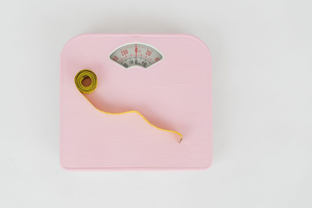 Pink analog bathroom scale and yellow measuring tape viewed from above, tools for monitoring weight management progress.