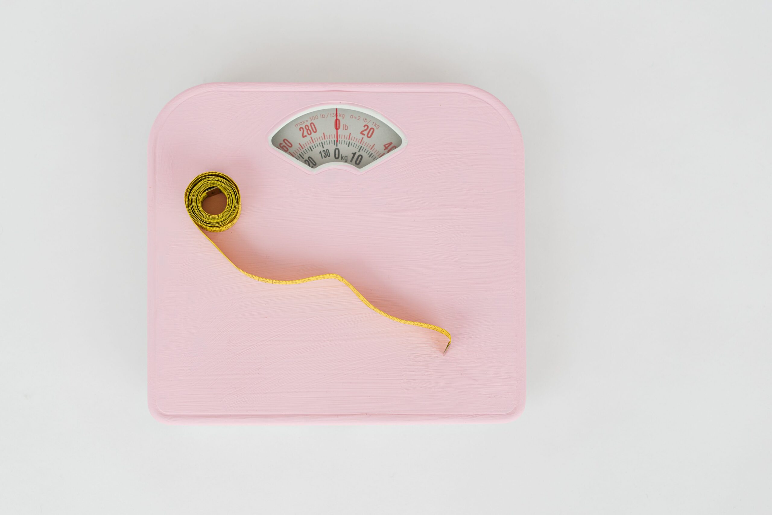 Pink analog bathroom scale and yellow measuring tape viewed from above, tools for monitoring weight management progress.