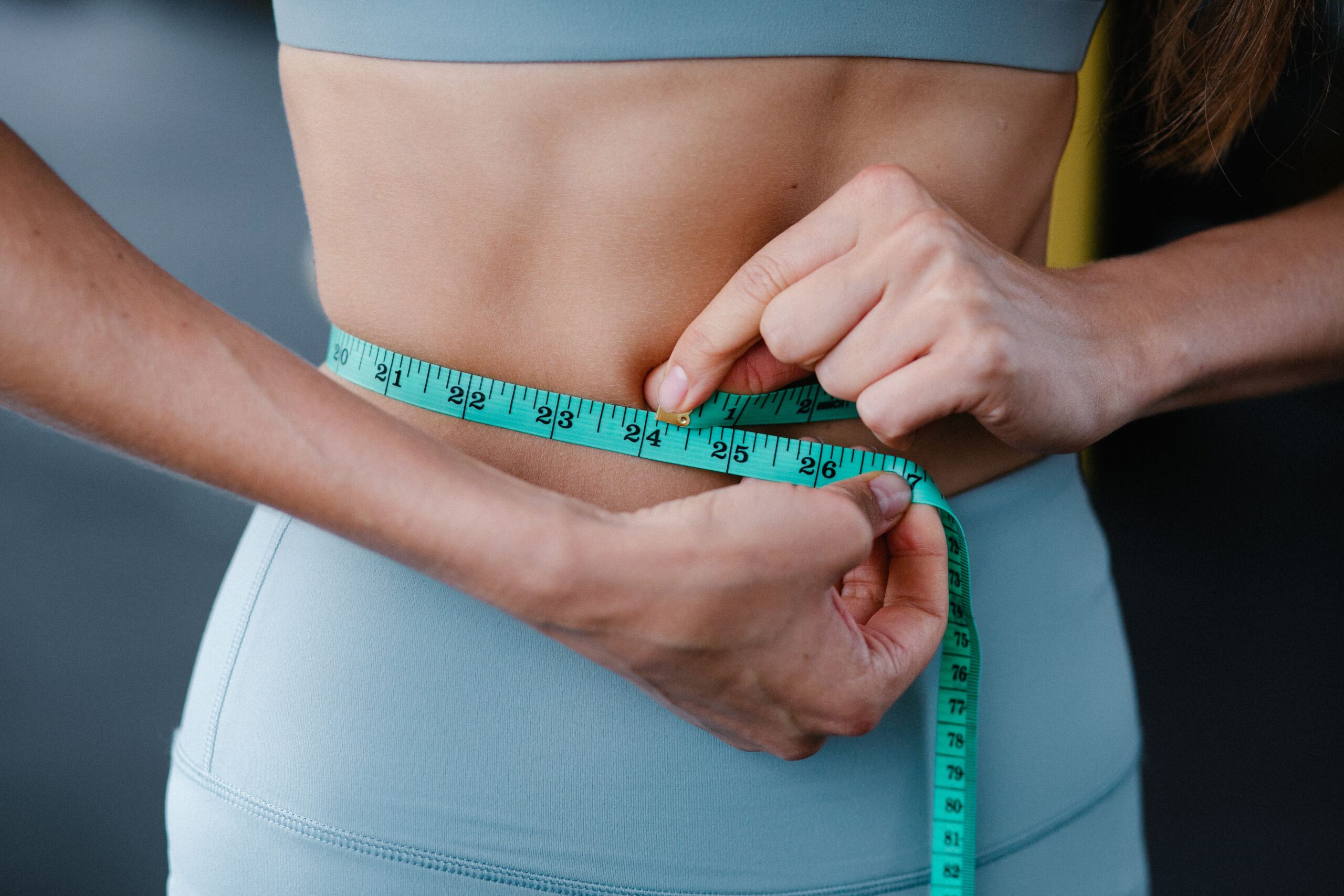 Woman in athletic wear measuring her waist with a green tape measure, tracking results while taking weight loss medications.