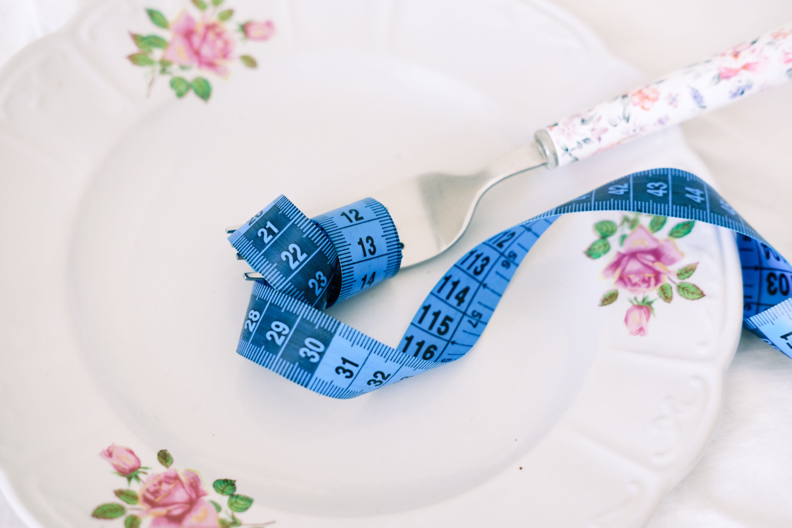 Blue measuring tape curled around a fork on an empty floral plate, symbolising reduced appetite from weight loss medications.