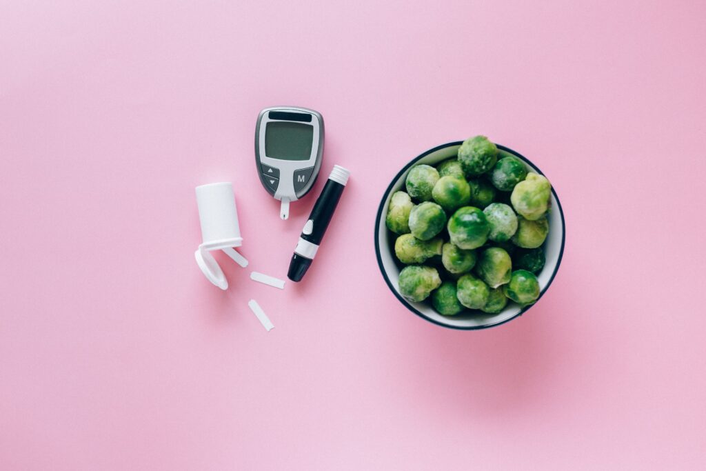 Blood glucose meter, lancet and test strips beside a bowl of Brussels sprouts on a pink background, illustrating diet monitoring alongside weight loss supplements.