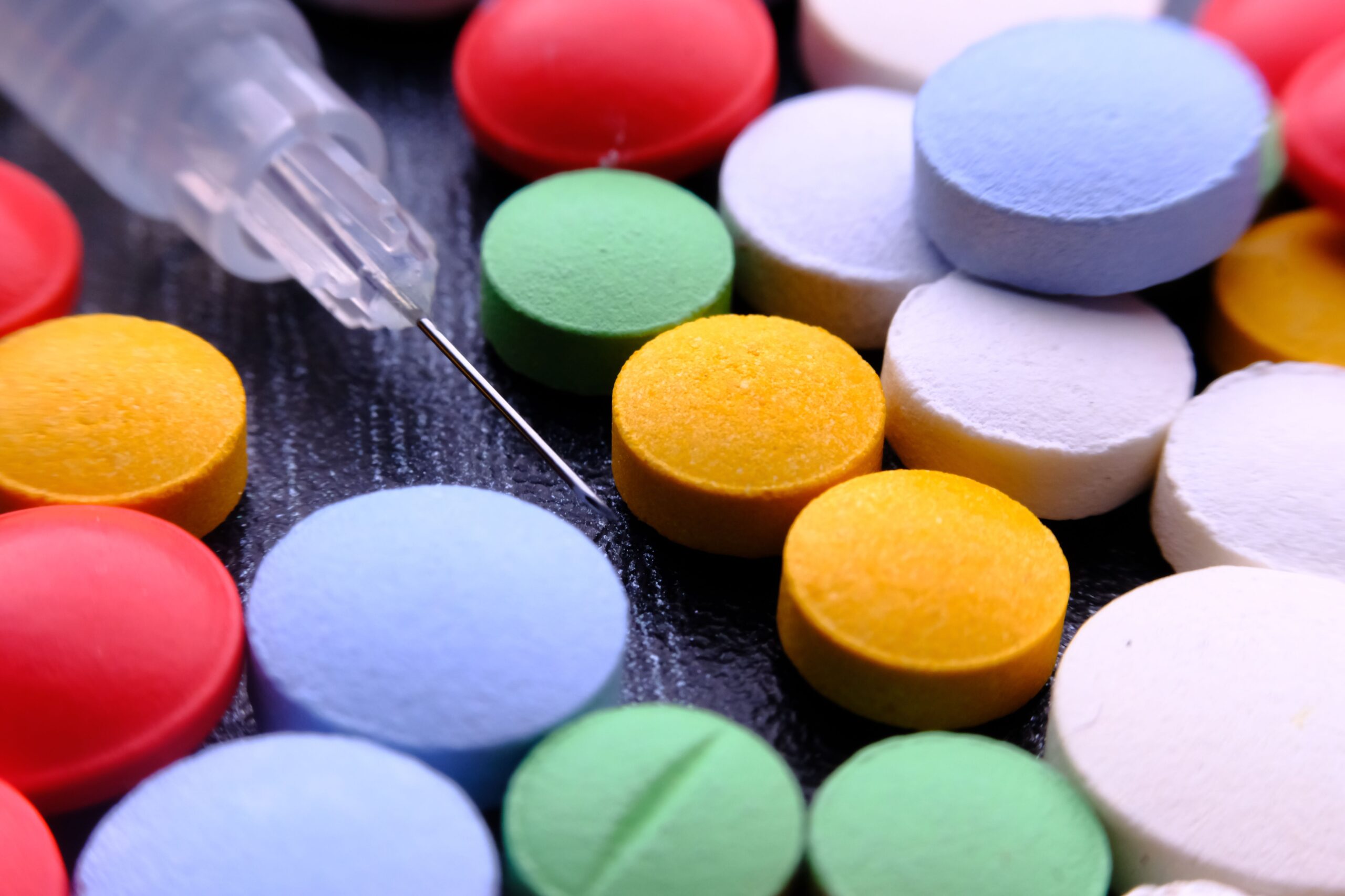 A weight loss medication syringe rests among scattered red, yellow, blue, green and white tablets on a dark surface.