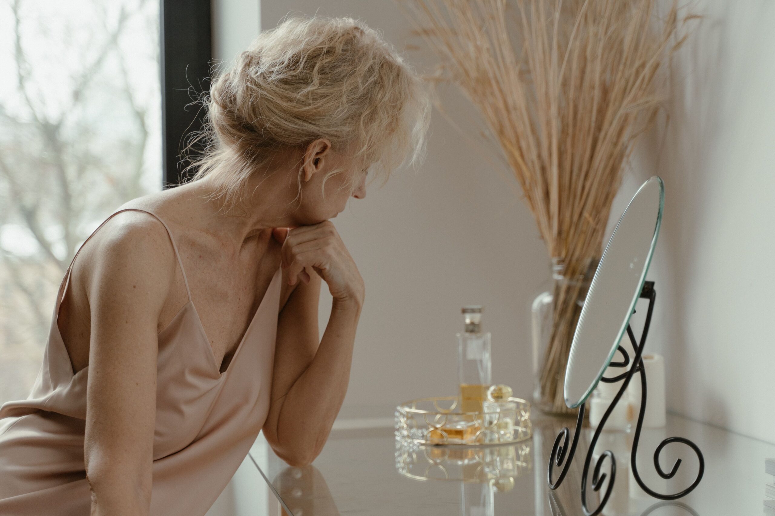 An older woman in a silk slip dress sits at a dressing table, head lowered and turned away from the mirror.