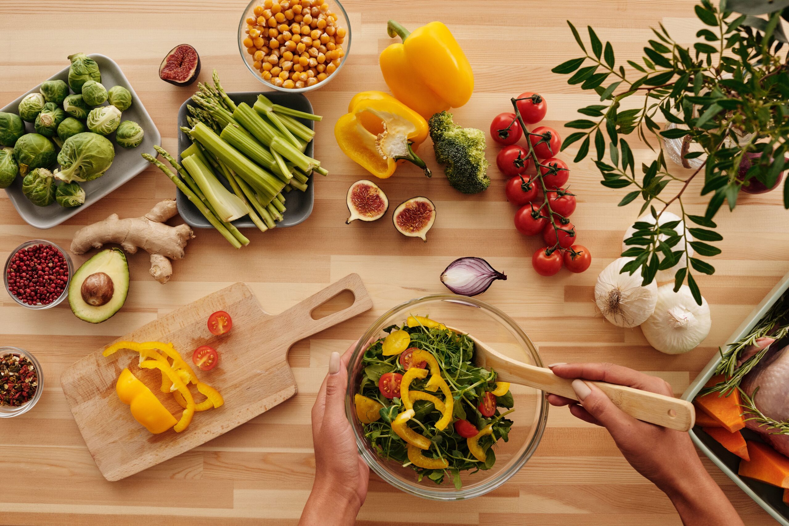 Overhead view of hands tossing a leafy salad, surrounded by fresh vegetables including sprouts, asparagus, peppers, tomatoes, avocado, garlic and chickpeas.