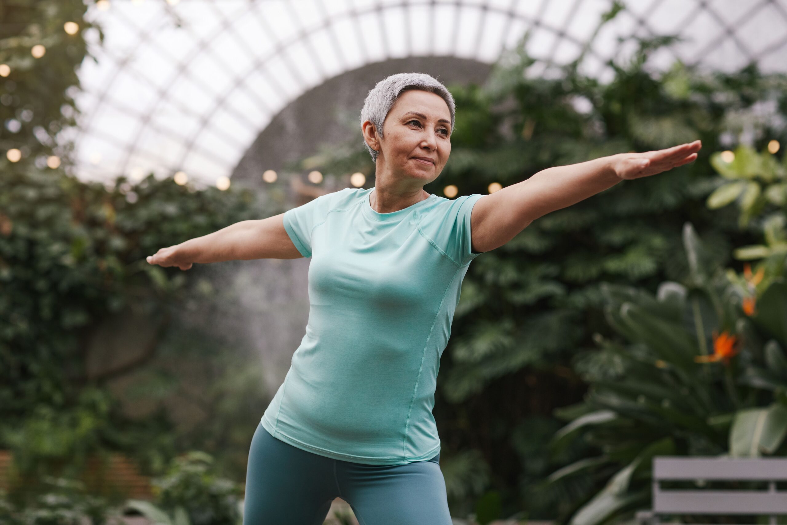 A grey-haired woman in a mint T-shirt holds a warrior yoga pose with arms outstretched, surrounded by greenery in a glasshouse.