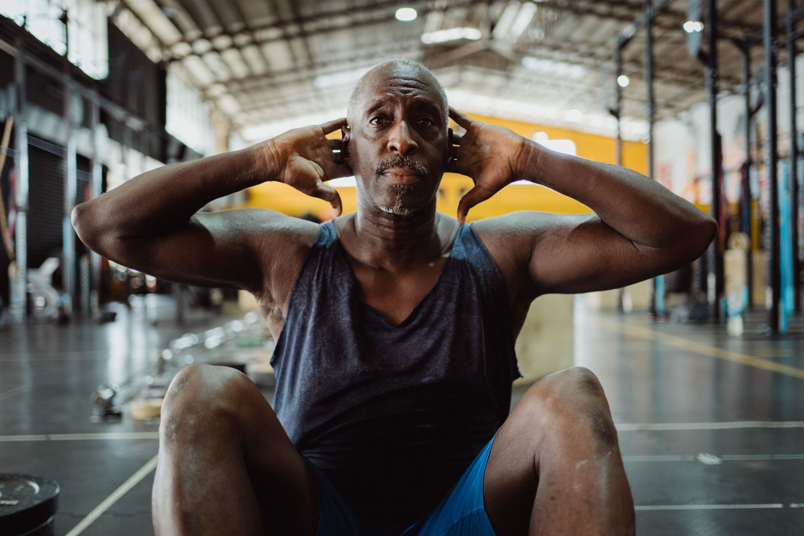 An older man in a vest and shorts performs sit-ups on the floor of an industrial-style gym, hands behind his head.