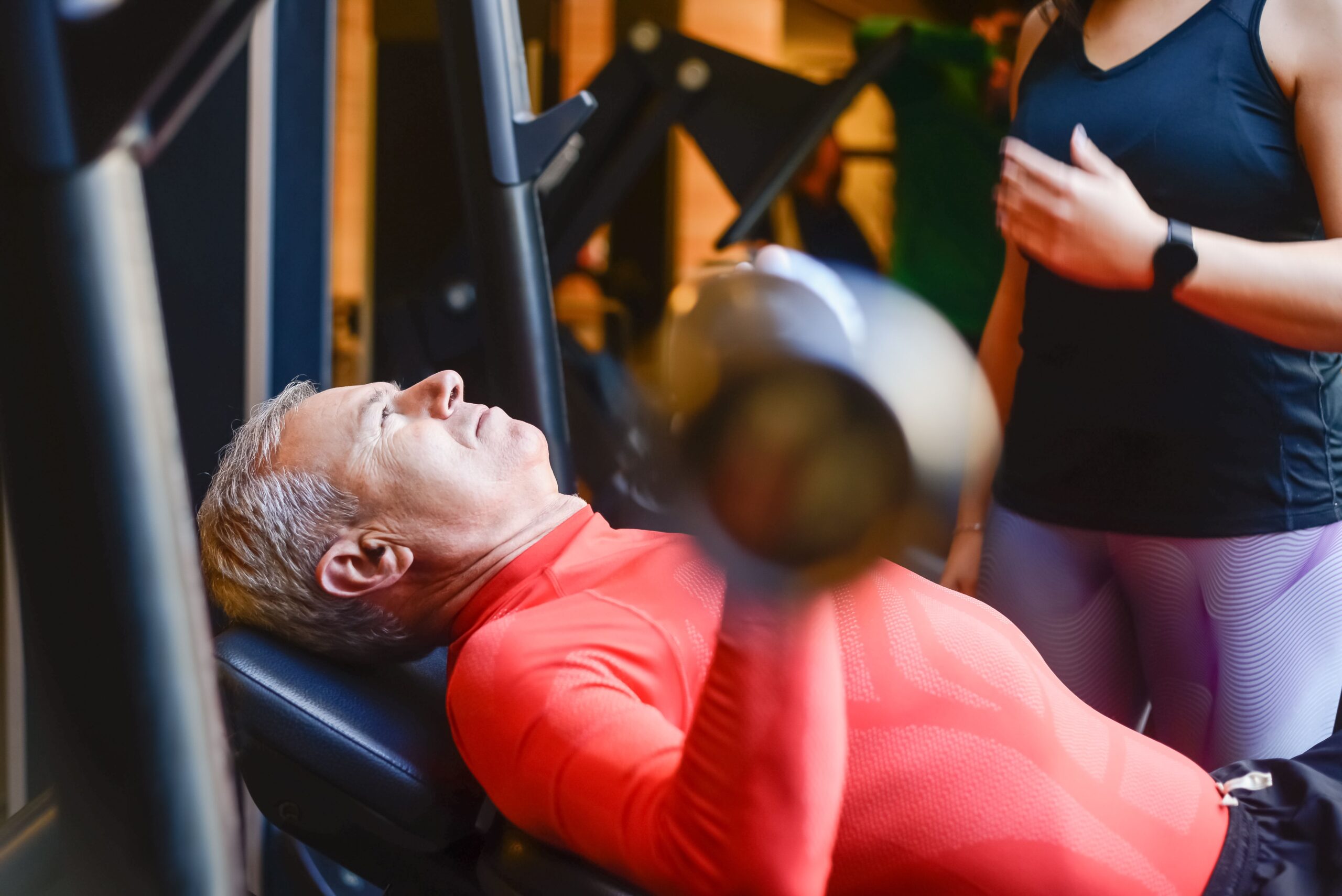 An older man in a red top lies on a bench pressing a barbell while a trainer stands beside him, spotting the lift.